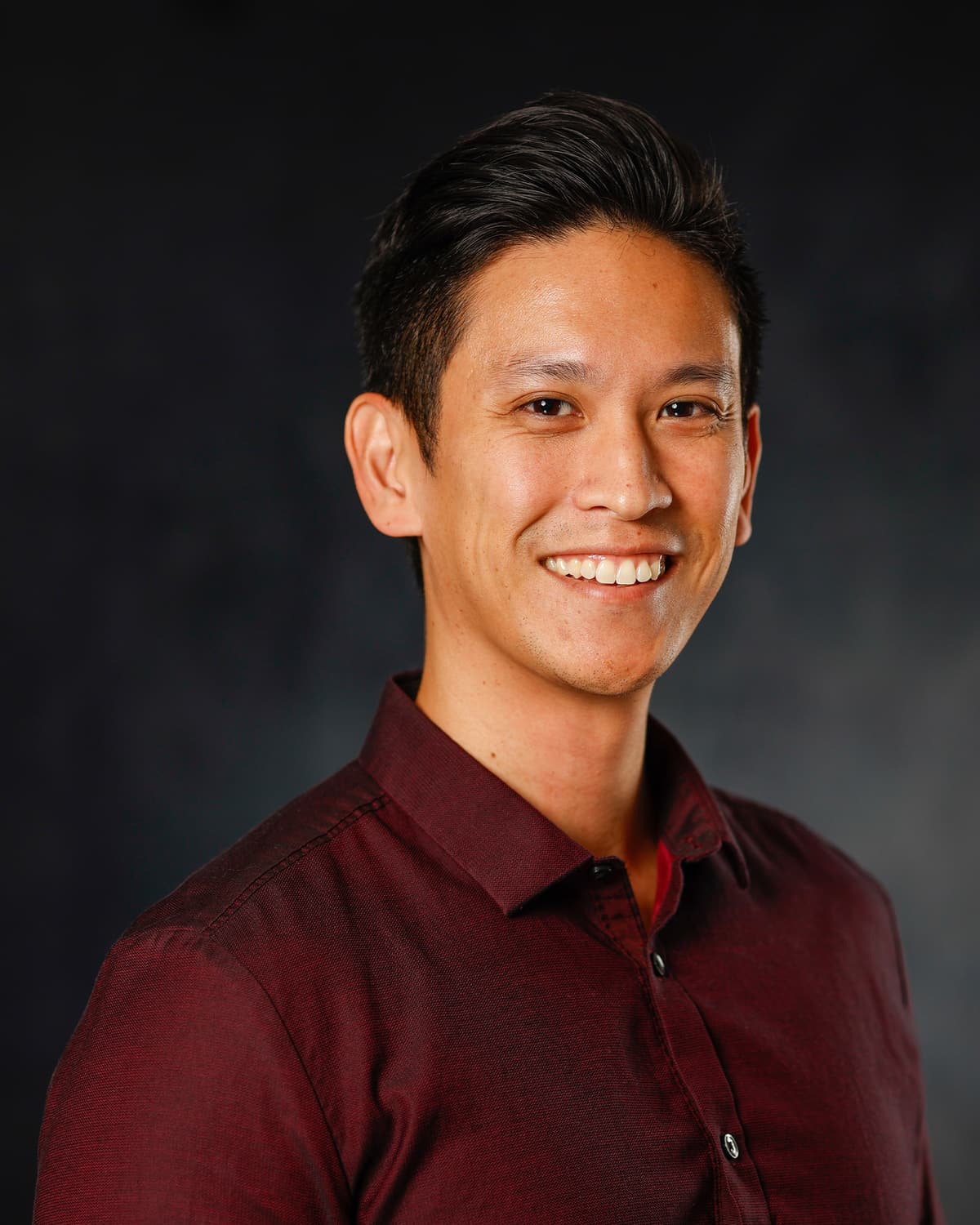 Smiling man in a burgundy button-up shirt in a studio headshot against a dark backdrop