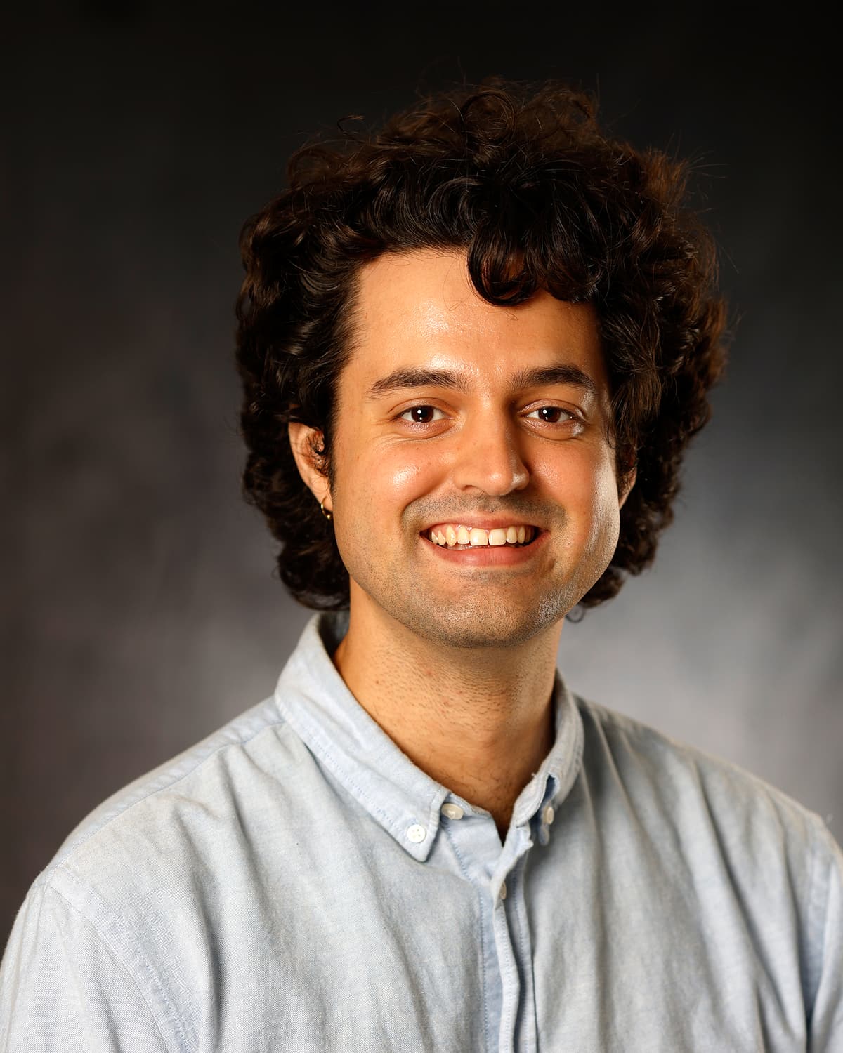 Smiling man with dark curly hair in a light blue oxford shirt in a studio headshot