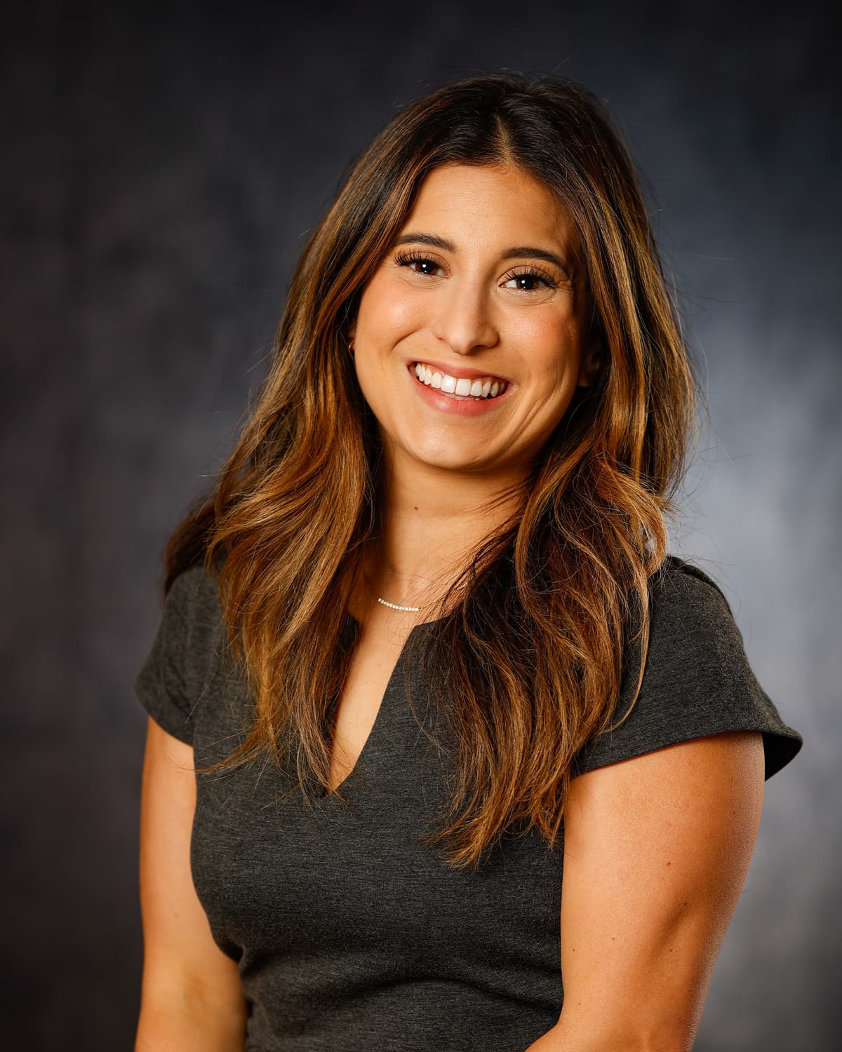 Smiling woman with long brown hair in a dark gray top in a studio headshot against a dark backdrop