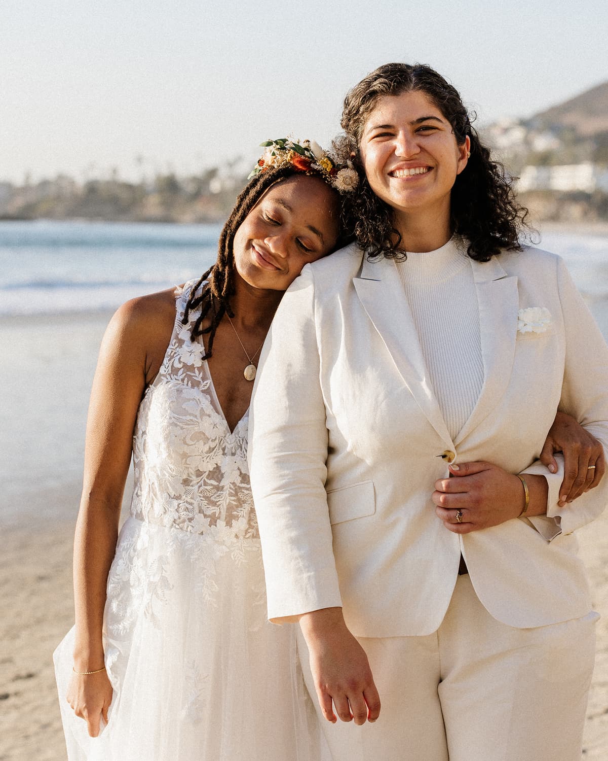 Two brides smiling together on the beach, one in a lace gown with a flower crown resting her head on the other, who wears a cream suit