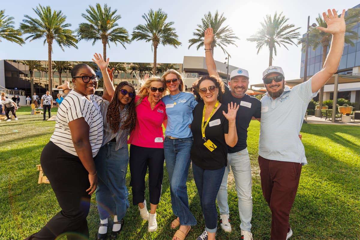 Group of colleagues cheering with raised arms at an outdoor company event on a sunlit lawn with palm trees