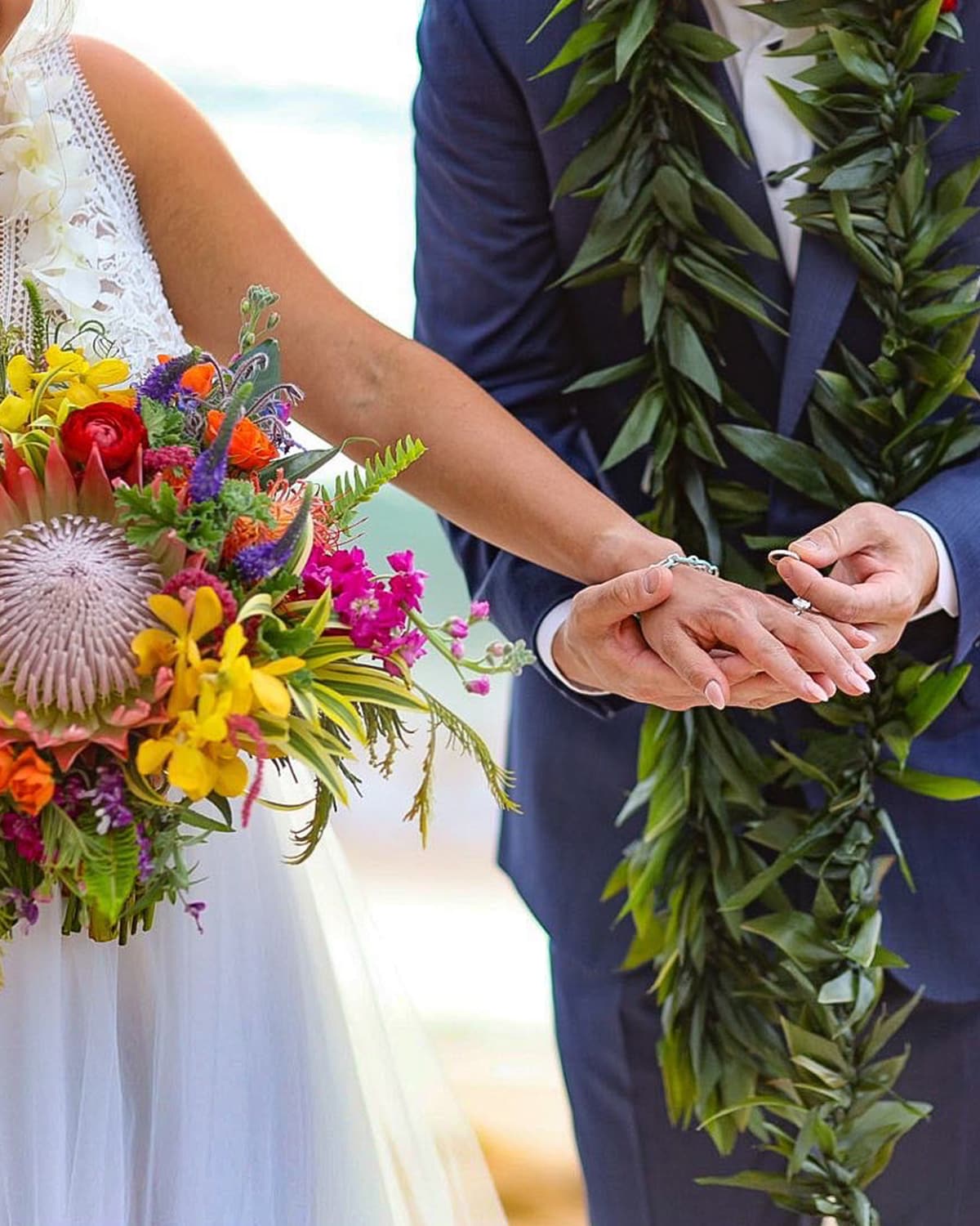 Couple exchanging rings at a beach ceremony, with a colorful tropical bouquet and a green leaf lei
