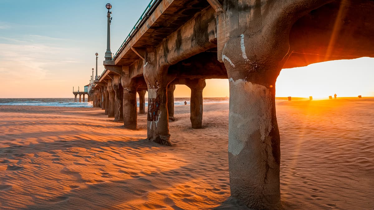 Beach pier at sunset with the sun flaring through weathered concrete pilings over rippled sand